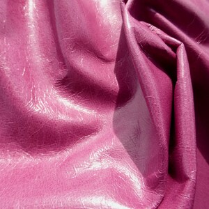 May include: Close-up of a deep pink, textured, faux leather fabric. The fabric is folded and draped, showing the texture and sheen.