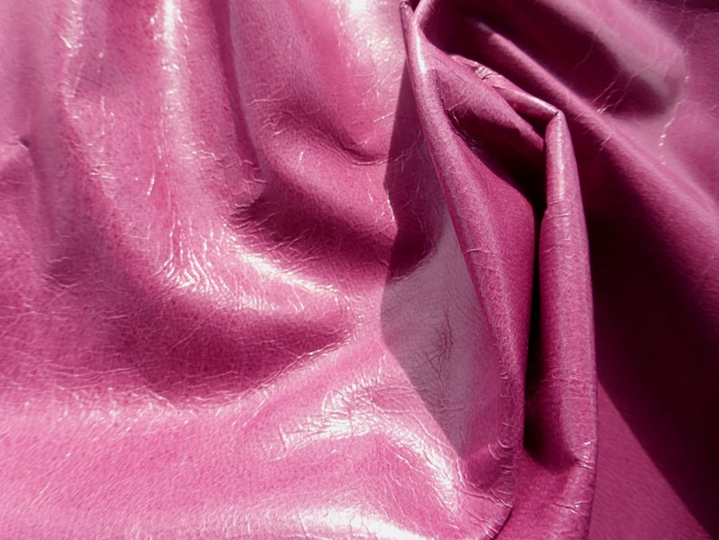 May include: Close-up of a deep pink, textured, faux leather fabric. The fabric is folded and draped, showing the texture and sheen.