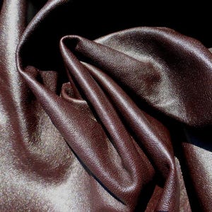 May include: Close-up of a dark brown leather material with a textured surface. The leather is folded and draped, creating shadows and highlights that emphasise its supple texture. The colour is a rich, deep brown.