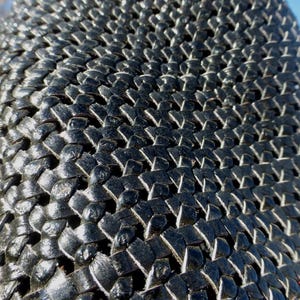 May include: Close-up of a section of black woven material. The weave pattern is tight and intricate, creating a textured surface. The material appears to be slightly glossy, reflecting light. The overall effect is a dark, geometric design.
