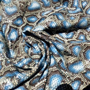 Pode incluir: Uma peça de material texturizado com um padrão repetitivo de escamas azuis e castanhas. A superfície apresenta um design distinto de escamas sobrepostas e em relevo que imita a textura da pele de réptil, criando profundidade visual.