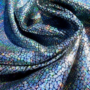 Op de afbeelding: Close-up van een iriserende blauwe en zilveren stof met een textuur van schubben. De stof is gevouwen en gedrapeerd, waardoor de glinsterende en reflecterende eigenschappen zichtbaar zijn.