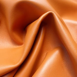 May include: Close-up of orange leather with visible texture and folds. The leather has a smooth, slightly shiny surface. The image highlights the material's flexibility and rich color.