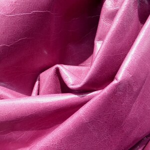 May include: Close-up of a deep pink leather fabric with a textured surface. The fabric is folded and draped, creating soft folds and curves.