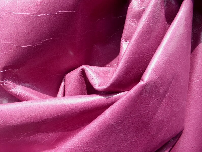 May include: Close-up of a deep pink leather fabric with a textured surface. The fabric is folded and draped, creating soft folds and curves.