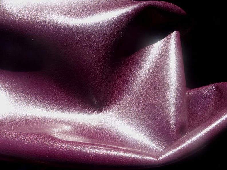 May include: Close-up of a deep purple leather fabric with a smooth, shiny surface. The fabric is folded and draped, creating soft folds and curves.