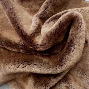 May include: A close-up of a brown faux fur fabric with dark brown accents. The fabric is soft and plush, with a swirled pattern. The material is likely used for clothing, blankets, or home decor.