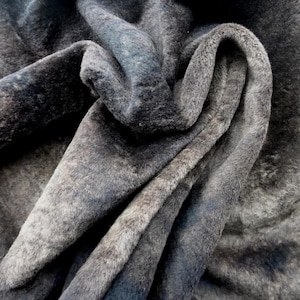 Könnte beinhalten: Nahaufnahme eines gefalteten, strukturierten Stoffes in Grau- und Schwarztönen. Das Material hat ein weiches, fellartiges Aussehen mit sichtbaren Falten und Schatten, die ein Gefühl von Tiefe und Textur erzeugen. Der Stoff wirkt weich und luxuriös.