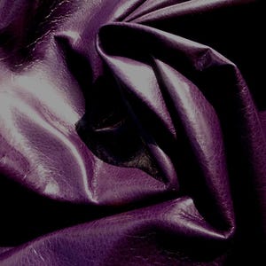 May include: Close-up of a deep purple leather fabric with a textured surface. The fabric is folded and draped, creating soft folds and curves.