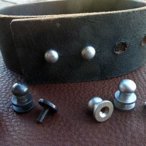 May include: A black leather cuff bracelet with silver-colored rivets. Several silver-colored screw-in rivets are displayed next to the bracelet. The bracelet is approximately 1.5 inches wide.