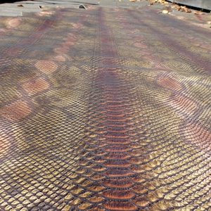 May include: A large piece of snakeskin leather with a scale pattern. The leather has a gradient of colours, including gold, brown, and reddish-brown. The scales are detailed and create a textured surface. The snakeskin is likely used for crafting.
