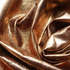 May include: Close-up of a textured, metallic gold fabric with folds and creases. The material has a shiny, reflective surface, suggesting it could be leather or a similar material. The fabric is likely used for fashion or crafting.