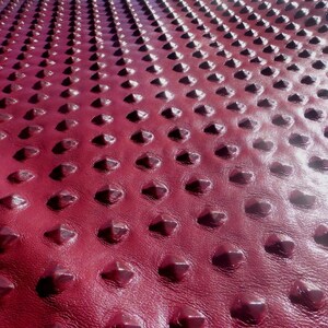 May include: Close-up of a deep red leather material with a textured, diamond-shaped pattern. The surface has a glossy finish.