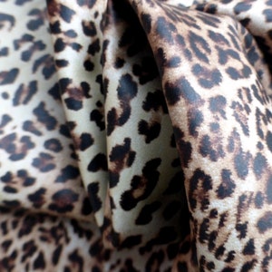 May include: Close-up of a brown and cream leopard print fabric with a satin finish. The fabric is folded and draped, showing the texture and pattern.