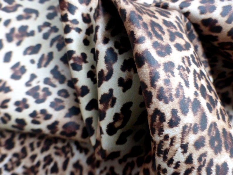 May include: Close-up of a brown and cream leopard print fabric with a satin finish. The fabric is folded and draped, showing the texture and pattern.
