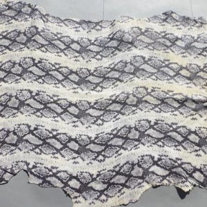 May include: A large, irregularly shaped piece of animal hide featuring a repeating snakeskin pattern in shades of charcoal gray, black, and off-white. The texture appears organic and flexible, with a distinct scale-like print across the surface.