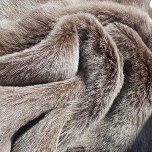 May include: A thick, plush faux fur fabric in a warm taupe and brown colour palette. The material features a dense, soft texture with long, layered fibres that create deep folds and shadows, highlighting the multi-tonal brown and greyish-tan hues.