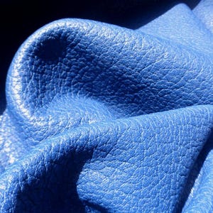 May include: A vibrant royal blue leather material with a distinct pebbled texture. The surface features deep folds and creases that create a play of light and shadow, highlighting the natural grain and flexible quality of the blue hide.