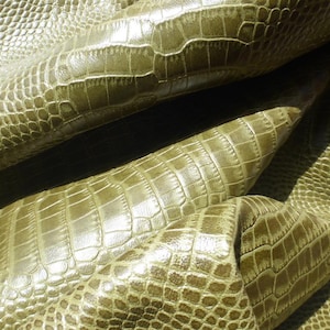 May include: A sheet of olive green leather featuring a raised, embossed crocodile skin texture. The material has a glossy, reflective finish with deep folds and creases that highlight the intricate, repeating scale pattern across the surface.