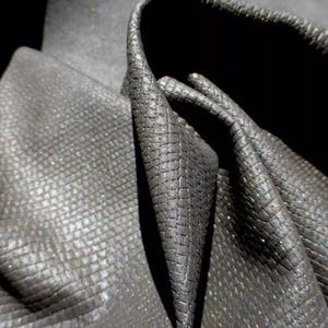 May include: Close-up of a dark gray fabric with a subtle shimmer. The fabric has a textured, almost scaly, surface.