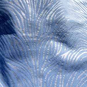 Op de afbeelding: Close-up van een blauwe stof met een zilveren metallic patroon. Het patroon lijkt op een reeks gebogen lijnen of golven.