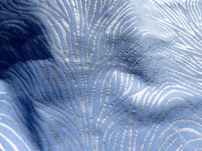 Op de afbeelding: Close-up van een blauwe stof met een zilveren metallic patroon. Het patroon lijkt op een reeks gebogen lijnen of golven.