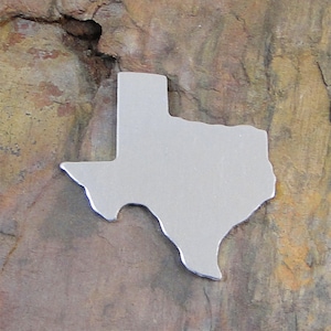May include: A silver metal cutout of the state of Texas. The cutout is on a brown, textured surface.