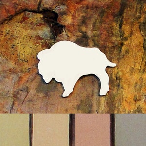 May include: A white silhouette of a buffalo on a brown and orange textured background. Below the buffalo is a row of four eyeshadow samples in shades of brown, beige, and gray.