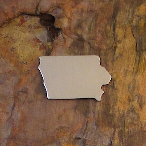 May include: A silver metal cutout of the state of Iowa. The cutout is on a brown, textured surface.