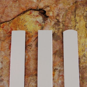 May include: Three white wooden slats against a textured, multi-colored background. Two slats have flat tops, while the third has a rounded top. The slats are evenly spaced and vertically oriented.