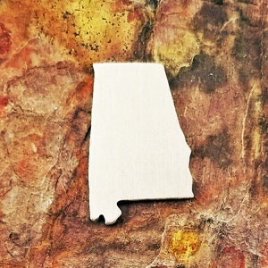 May include: A wooden cutout of the state of Alabama, USA, is centered against a textured, multi-colored background of browns, reds, and yellows. The cutout is a light, natural wood color.
