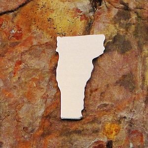 May include: A white metal cutout of the state of Vermont. The cutout is on a brown, textured surface.