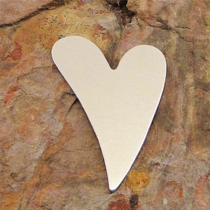 May include: A silver heart-shaped metal charm, with a smooth, polished surface. The charm is sitting on a textured, brown rock.