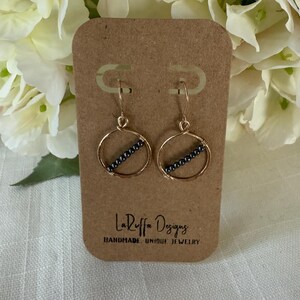 May include: Gold-tone hoop earrings with a diagonal row of small, dark gray beads. The earrings are displayed on a brown card with the text "LaRuffa Designs HANDMADE UNIQUE JEWELRY". The background features white flowers.