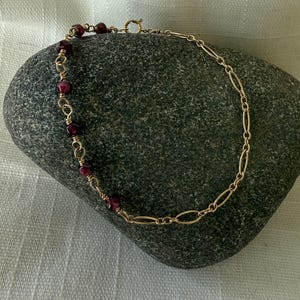 May include: A gold chain bracelet with small, dark red beads. The bracelet is on a gray rock.