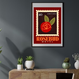 May include: Framed art print with a rose design in a postage stamp style. The print features the text "SCHITT'S CREEK" and "ROSEBUD MOTEL" in orange against a dark red background. The artwork is displayed on a wooden cabinet, with plants in the foreground.