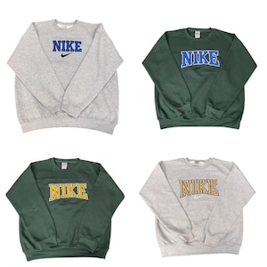 Pu&ograve; includere: Quattro felpe girocollo Nike. Due sono grigie, una con scritta "NIKE" blu e una dorata. Le altre due sono verdi, una con scritta "NIKE" blu e una dorata. Tutte presentano il logo Nike.