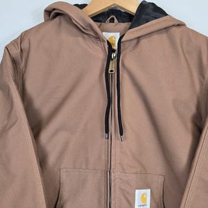 May include: A brown hooded Carhartt jacket with a black zipper and a black drawstring. The jacket has a Carhartt logo patch on the left chest.