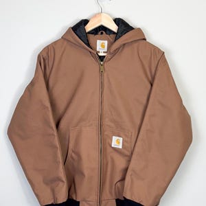 May include: A brown Carhartt hooded jacket with a black zipper and black trim. The jacket has a Carhartt logo patch on the left chest.