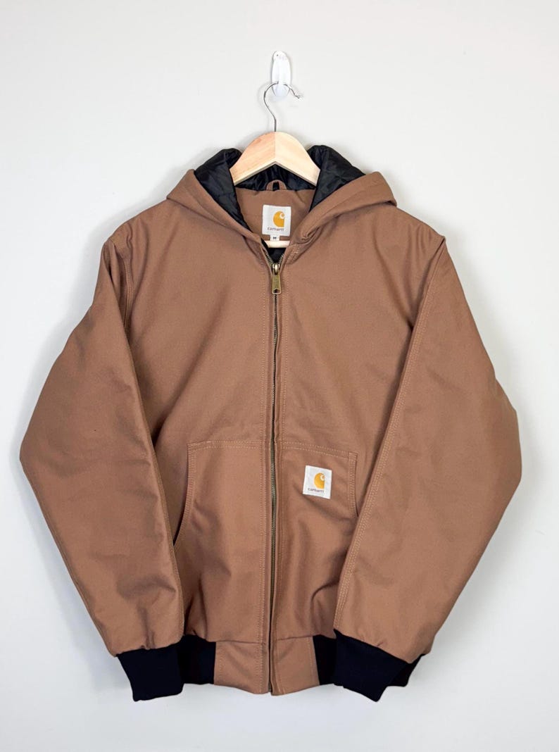 May include: A brown Carhartt hooded jacket with a black zipper and black trim. The jacket has a Carhartt logo patch on the left chest.
