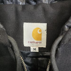 May include: Black Carhartt hooded jacket with a zipper closure. The jacket has a label with the Carhartt logo and the size "M" printed on it.