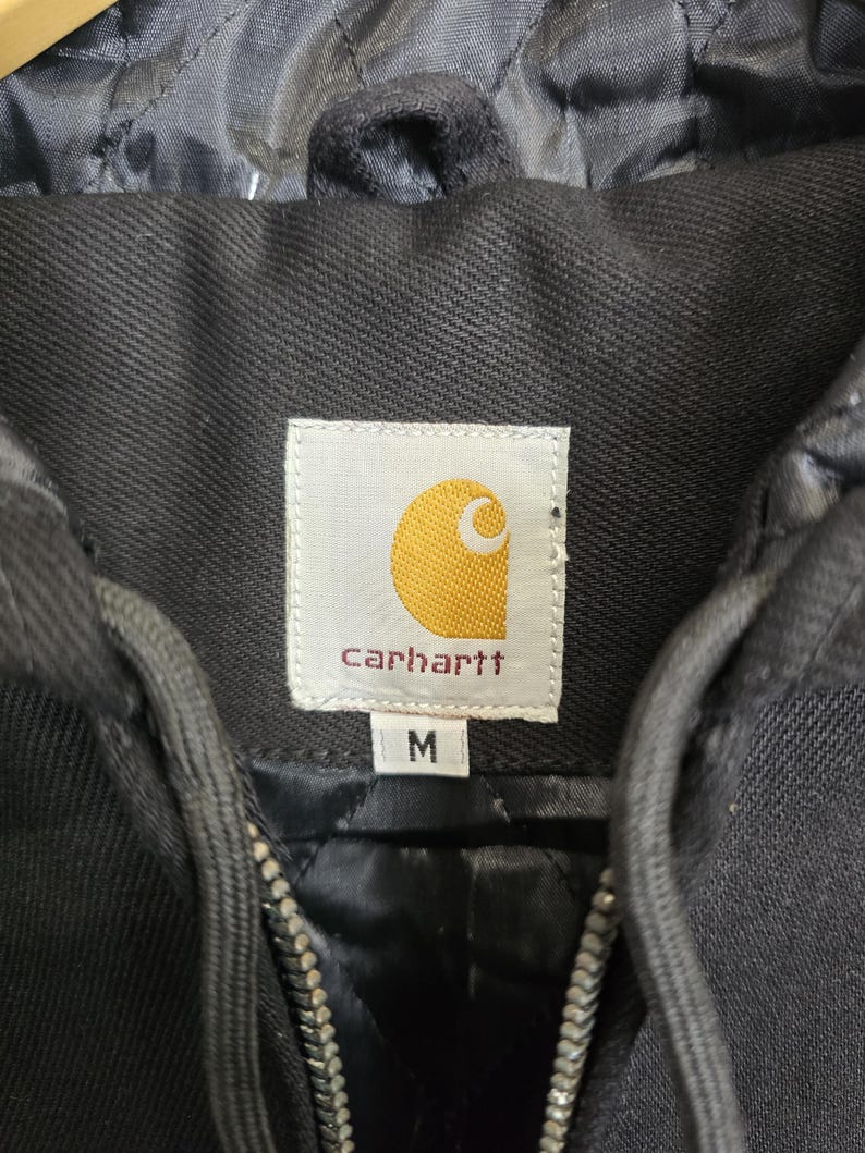 May include: Black Carhartt hooded jacket with a zipper closure. The jacket has a label with the Carhartt logo and the size "M" printed on it.