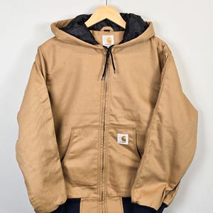 Op de afbeelding: Een bruin-beige jas met capuchon, zwarte gewatteerde voering en een ritssluiting aan de voorkant. De jas heeft een klein vierkant logopatch op de voorzak en donkerblauwe geribde manchetten. Op een houten hanger.