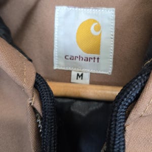 May include: A brown Carhartt jacket with a black zipper and black drawstrings. The jacket has a label that says "Carhartt" and "M" for size.