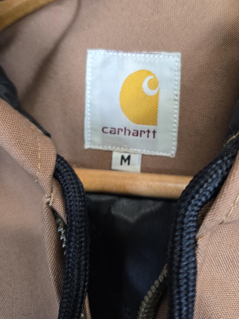 May include: A brown Carhartt jacket with a black zipper and black drawstrings. The jacket has a label that says "Carhartt" and "M" for size.