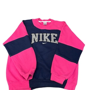 Op de afbeelding: Een sweatshirt met lange mouwen en ronde hals met een color-block ontwerp in felroze en marineblauw. In het midden van de borst staat het woord NIKE in grote grijze blokletters met een klein wit swoosh-logo direct daaronder.