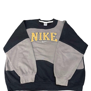 Op de afbeelding: Een sweatshirt met lange mouwen en ronde hals met een kleurgeblokt ontwerp in zwart en grijs. Op de borst staat het woord NIKE in gele blokletters met een zwarte omlijning, geplaatst boven een klein wit geborduurd swoosh-logo.