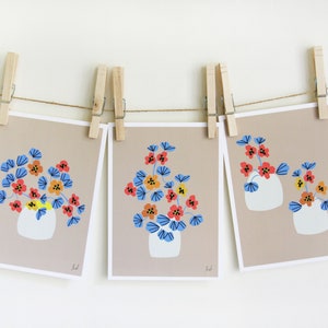 Op de afbeelding: Drie kunstdrukken met een bloemmotief. Elke print toont een witte vaas gevuld met rode, gele en oranje bloemen met blauwe bladeren tegen een beige achtergrond. De prints zijn met houten wasknijpers aan een touwtje bevestigd.