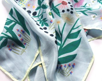May include: A light blue silk scarf with a floral print featuring green leaves, pink, yellow, and white flowers, and black polka dots. The scarf has a light yellow border and the text "wildflowers" and "floral dreams" printed on it.