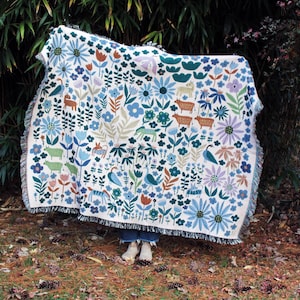Puede incluir: Una manta crema con un estampado floral y animal. El diseño presenta flores azules, blancas y marrones, junto con ilustraciones de pájaros, ciervos y vacas. La manta tiene un borde con flecos.